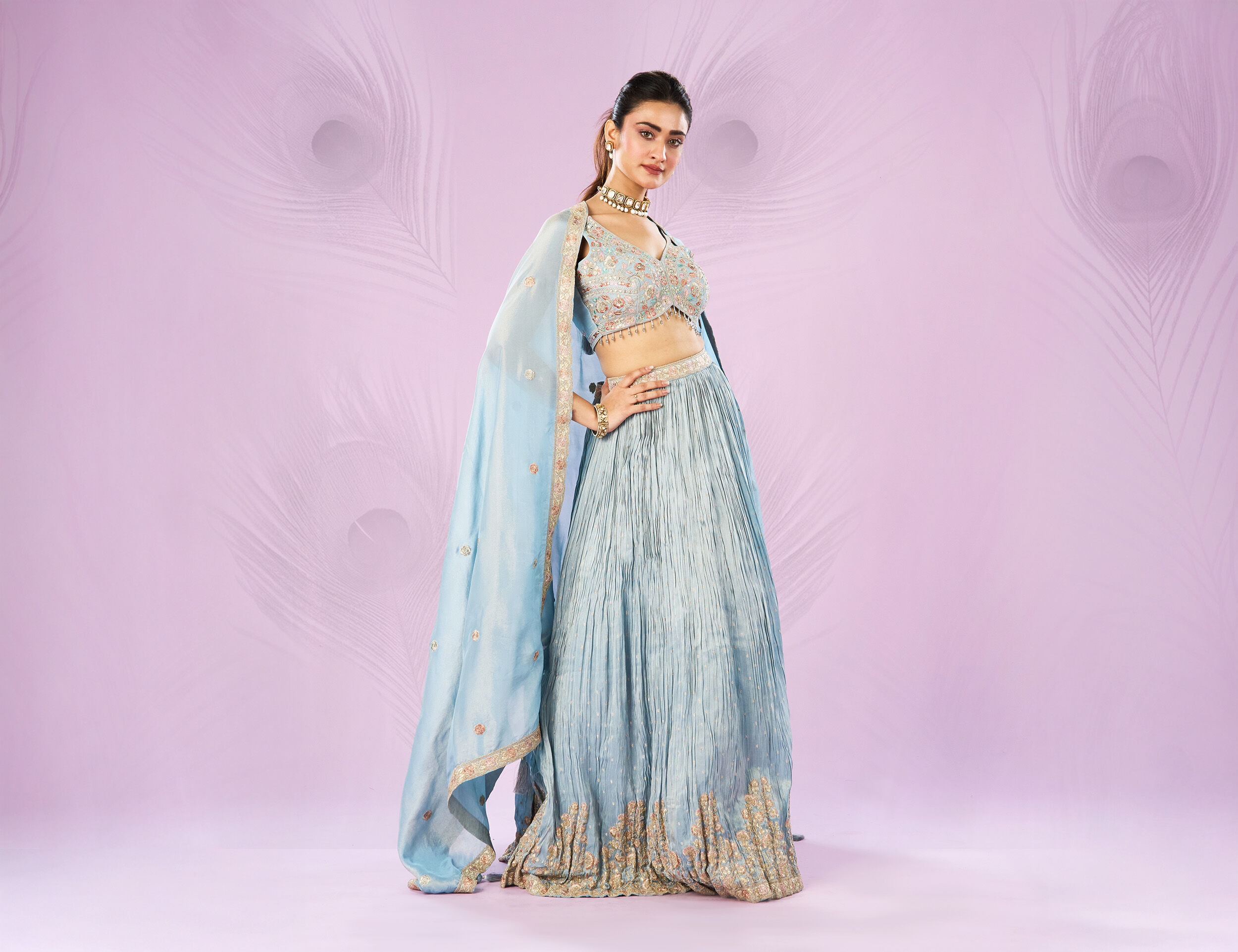 Woman wearing a pastel blue embroidered lehenga with matching dupatta, styled with traditional jewelry, posing against a soft pink background with peacock feather motifs.